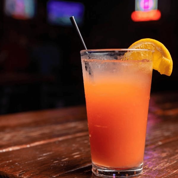 A vibrant orange cocktail with an orange slice and black straw on a dark wooden bar.