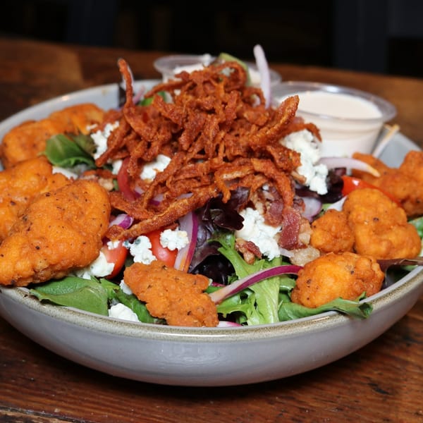 Crispy chicken salad with fried onions, bacon, feta, tomatoes, and red onion, with dressing cups.