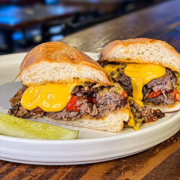 Two halves of a cheesy steak and pepper sub with a pickle spear on a white plate.