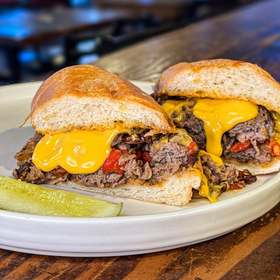 Two halves of a cheesy steak and pepper sub with a pickle spear on a white plate.