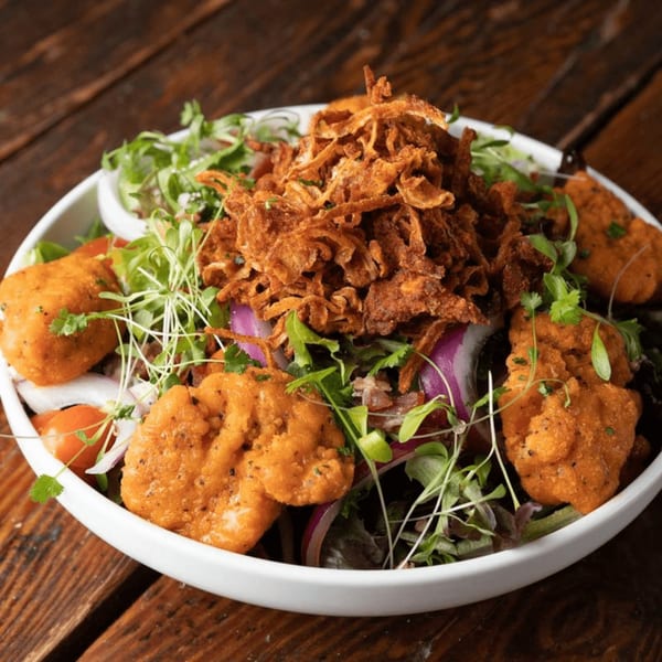 Crispy chicken salad with fried onions, red onion, tomatoes, and microgreens in a white bowl.