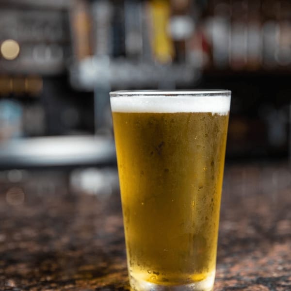 A frosty glass of golden beer with a foamy head sits on a dark bar counter.