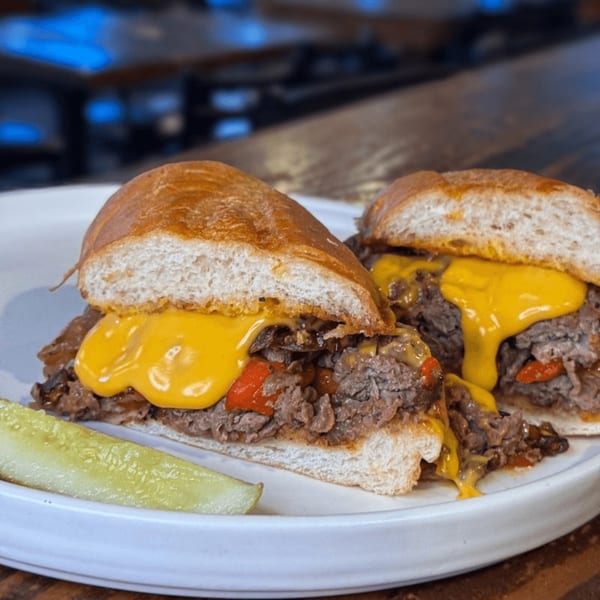 A Press Box Sports Grill cheesesteak sandwich with melted cheese, steak, peppers, and a pickle spear.